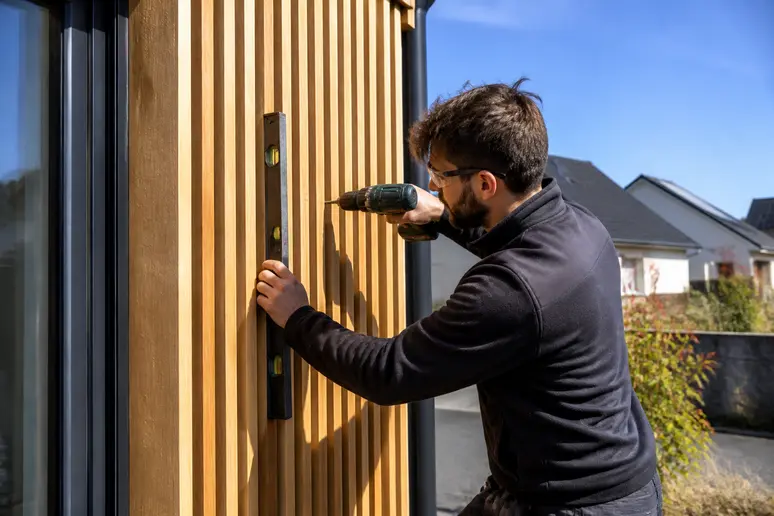 Un artisan français pose avec précision des lames de bardage en bois en claire-voie sur une façade, en plein soleil.