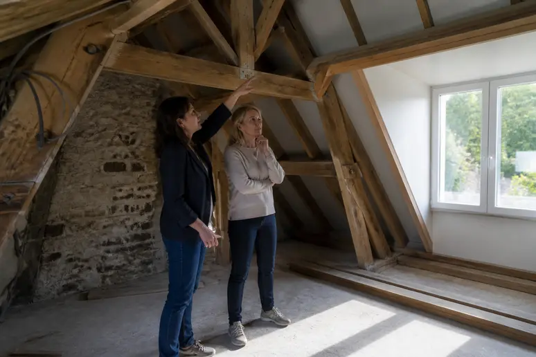Une architecte française présente un projet d'aménagement de combles à un propriétaire, illustrant les adaptations nécessaires.