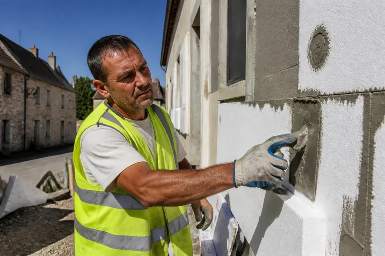 Un ouvrier français en gilet de sécurité pose des panneaux isolants sur un mur extérieur, dans une rue de village typique.