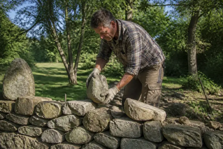Un homme pose méthodiquement des pierres pour ériger un mur en pierre sèche dans un jardin ensoleillé.