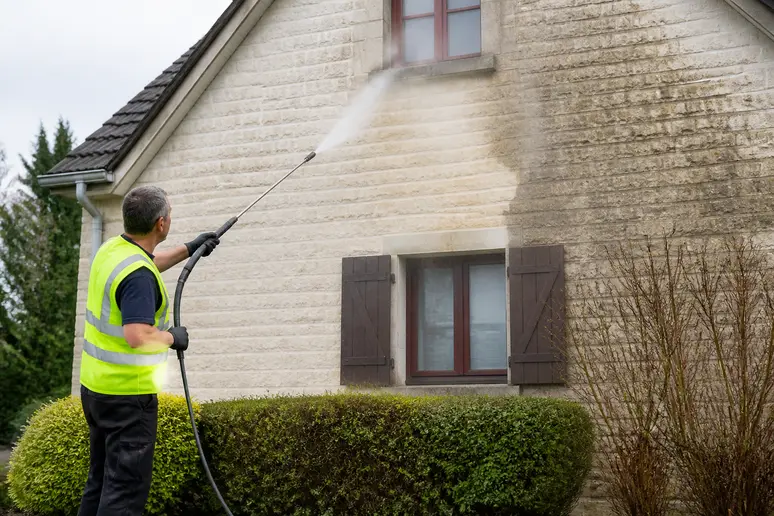 Un homme opere un nettoyeur haute pression sur la façade d'une maison en pierre, visant à enlever la saleté incrustée.