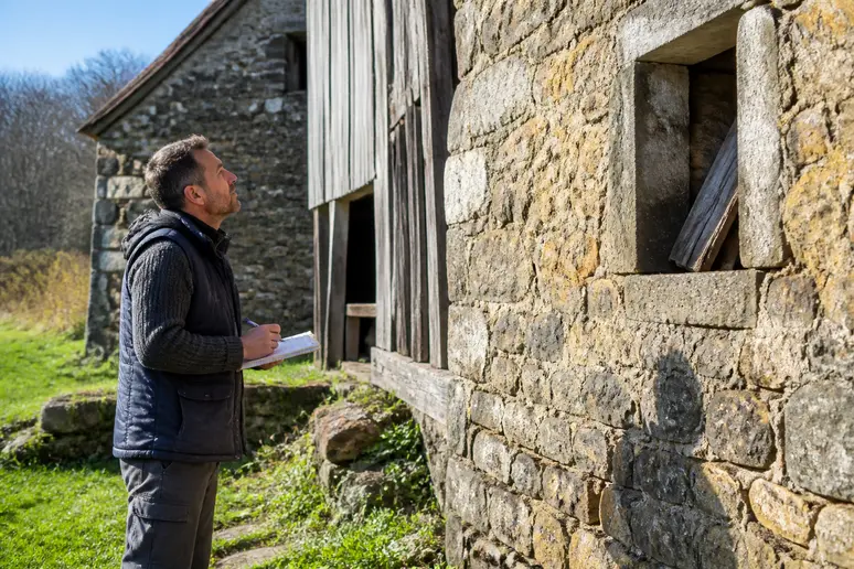 Un homme d'âge mûr examine avec attention une ancienne grange en pierre, prenant des notes dans un carnet.