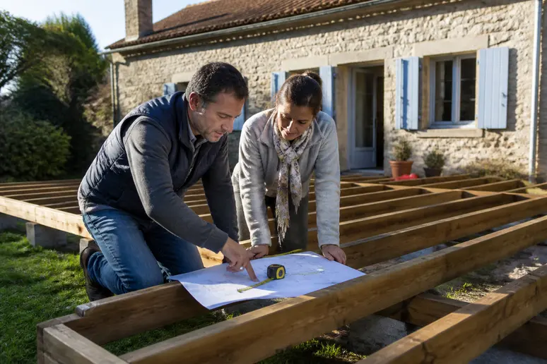 Couple français examinant les plans d'une terrasse en bois surélevée, en cours de construction près de leur maison.