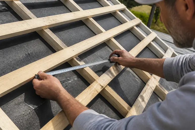 Artisan mesurant l'espacement des liteaux sur une charpente en bois pour poser des tuiles.